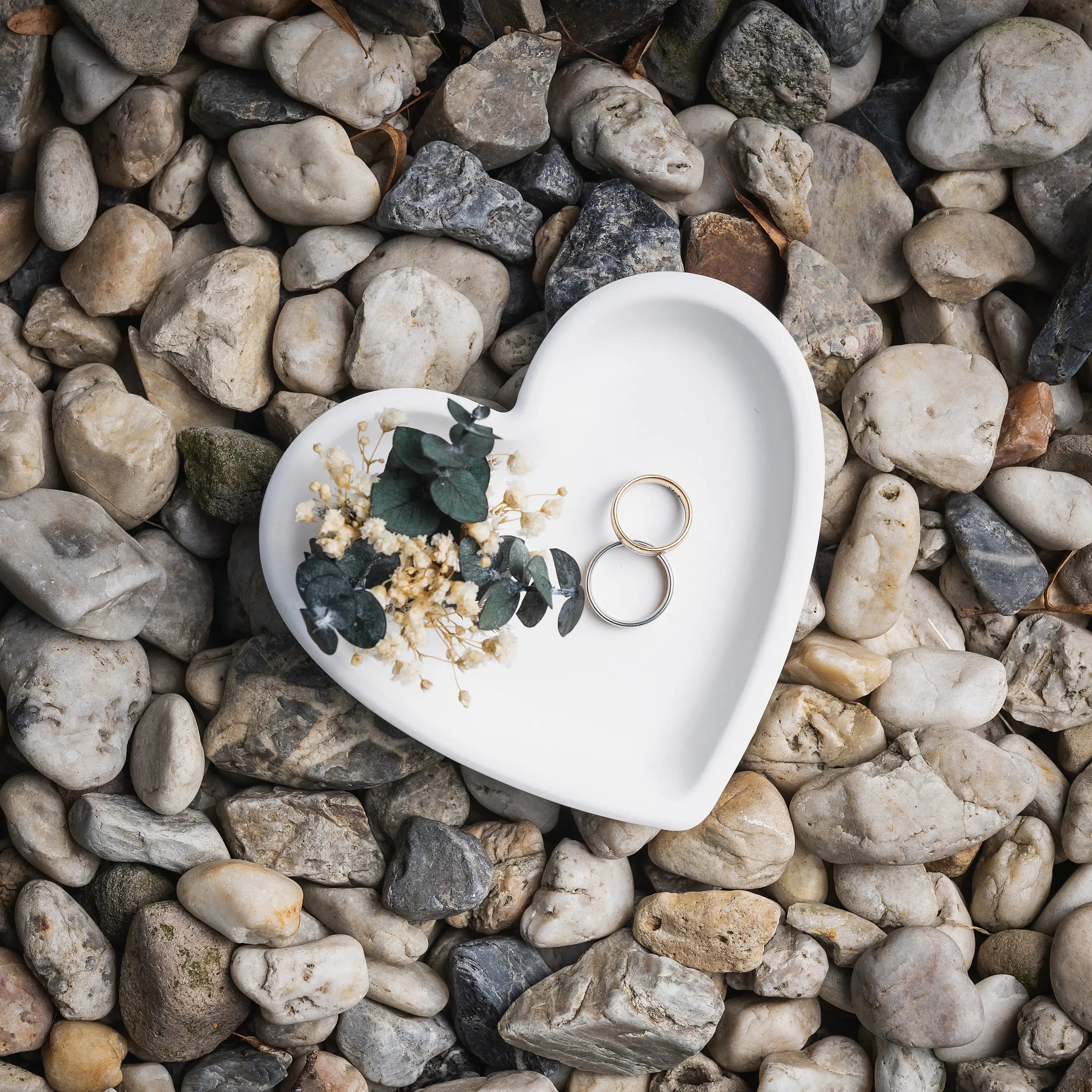 Herzformige Keramikschale mit Trockenblumen und Ringen auf Kieselsteinen